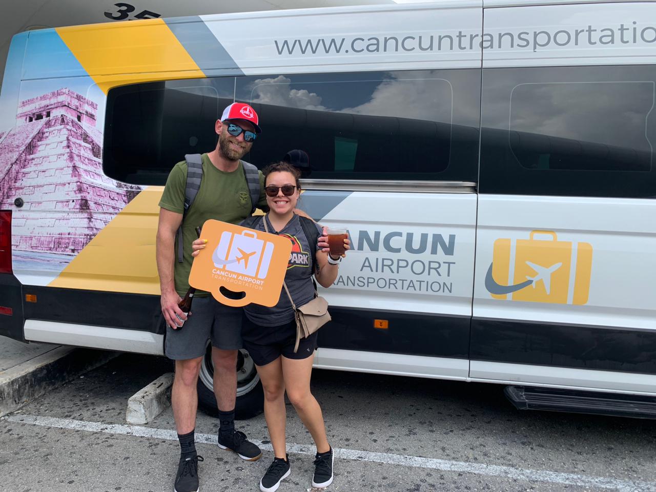 Happy travelers in sunglasses holding an orange luggage sign in front of a white-and-yellow Cancun airport shuttle van