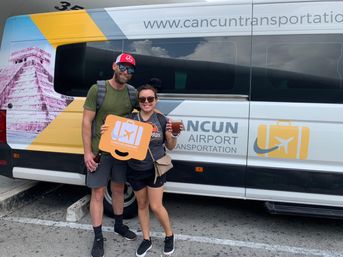 Happy travelers in sunglasses holding an orange luggage sign in front of a white-and-yellow Cancun airport shuttle van