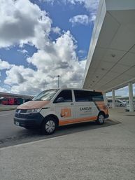 Orange-and-white airport shuttle van parked at a Cancun airport terminal curb under a bright blue sky with puffy clouds.
