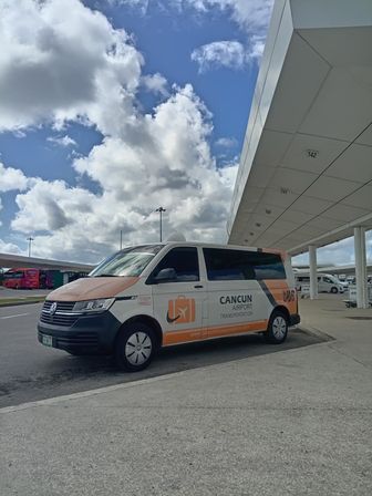 Orange-and-white airport shuttle van parked at a Cancun airport terminal curb under a bright blue sky with puffy clouds.