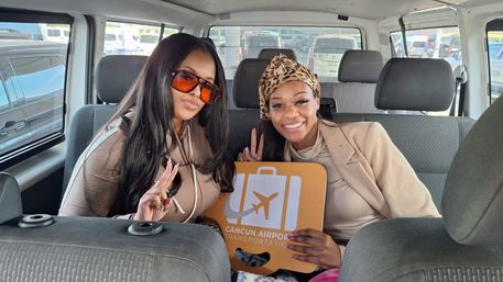 Two smiling women giving peace signs inside a shuttle van, holding an orange Cancun airport shuttle sign, wearing sunglasses, headscarf and casual beige outfits ready for travel.