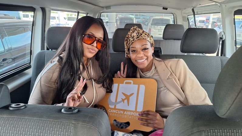 Two smiling women giving peace signs inside a shuttle van, holding an orange Cancun airport shuttle sign, wearing sunglasses, headscarf and casual beige outfits ready for travel.