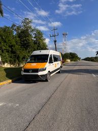 White-and-orange passenger shuttle van parked on a sunny tropical roadside curb with palm trees, power lines and a communications tower beside an empty highway under a blue sky.