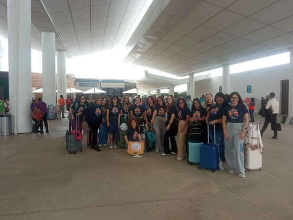 Group of travelers in matching navy T-shirts posing with colorful rolling suitcases and backpacks under a covered airport terminal curbside, ready for a group trip