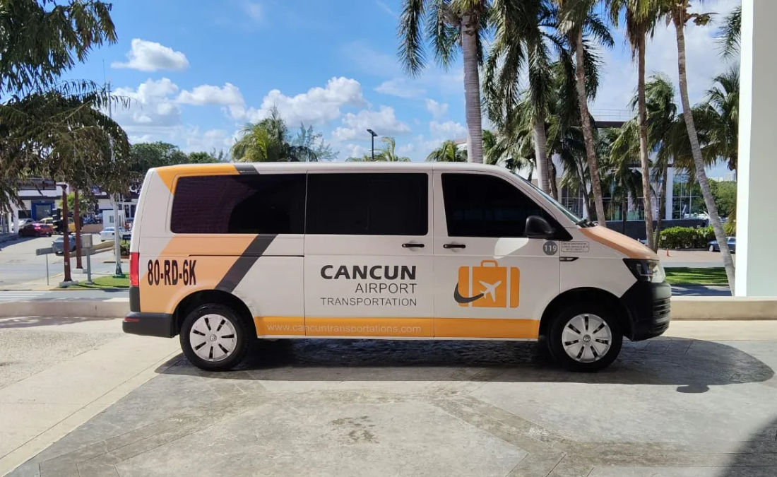White-and-orange airport shuttle van with a luggage icon parked under palm trees at Cancun airport pickup area on a sunny day
