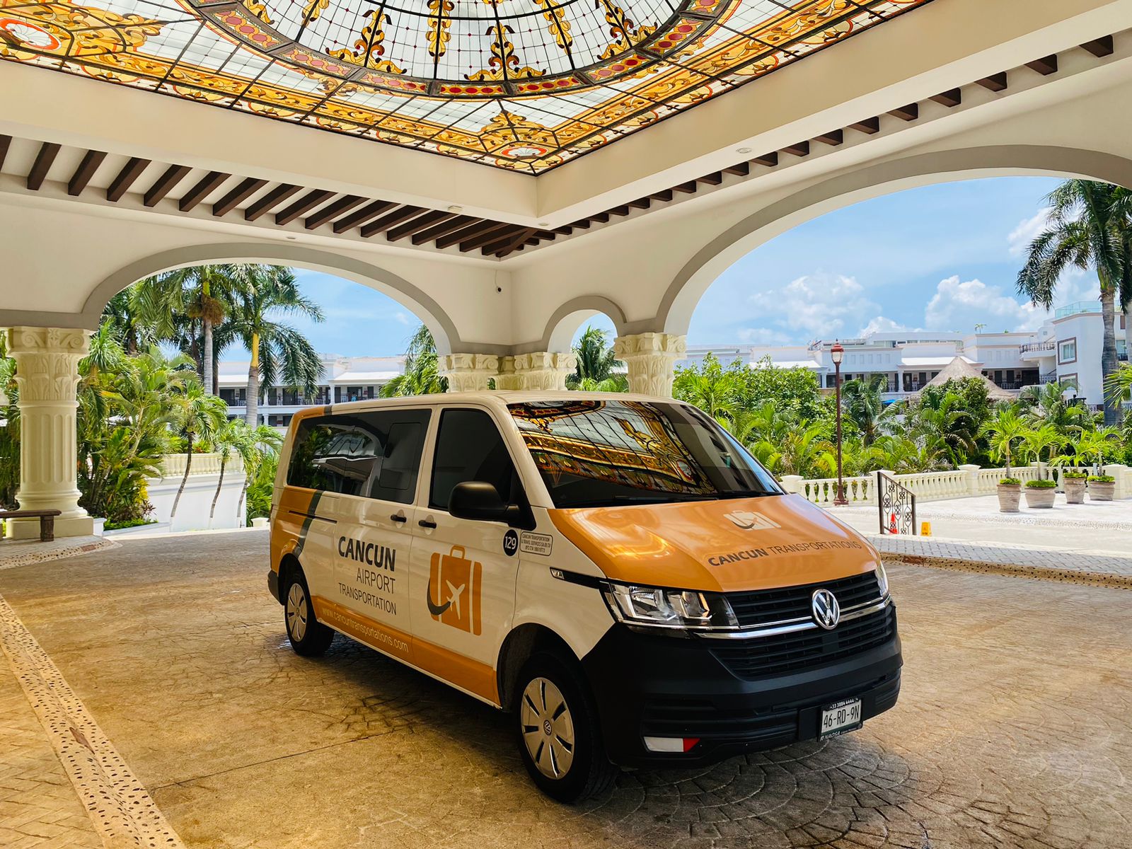 Orange-and-white airport shuttle van parked under an ornate stained-glass ceiling at a tropical Cancun resort entrance, with palm trees and blue sky in the background.