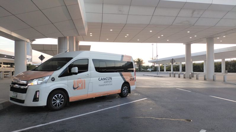 White-and-orange airport shuttle van parked under a modern terminal canopy at Cancun airport at sunrise, ready for passenger pickup.