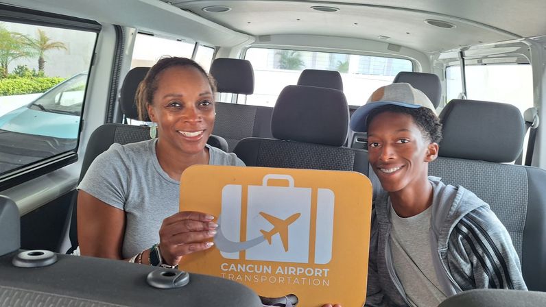 Smiling woman and teen in a shuttle van holding an orange airport shuttle sign for Cancun airport transfer.
