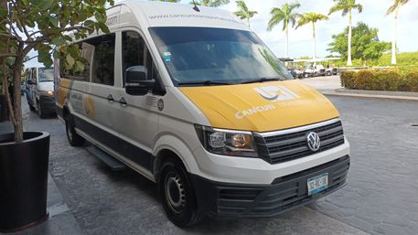 White-and-yellow airport shuttle van parked at a tropical Cancun resort drop-off with palm trees and golf carts in the background