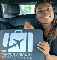 Smiling woman in a car holding a Cancún airport shuttle sign featuring a suitcase graphic and airplane icon