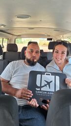 Two passengers sitting in a shuttle van smiling and holding an airport transfer sign for Cancun airport.