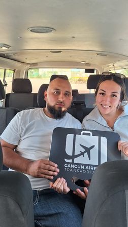 Two passengers sitting in a shuttle van smiling and holding an airport transfer sign for Cancun airport.