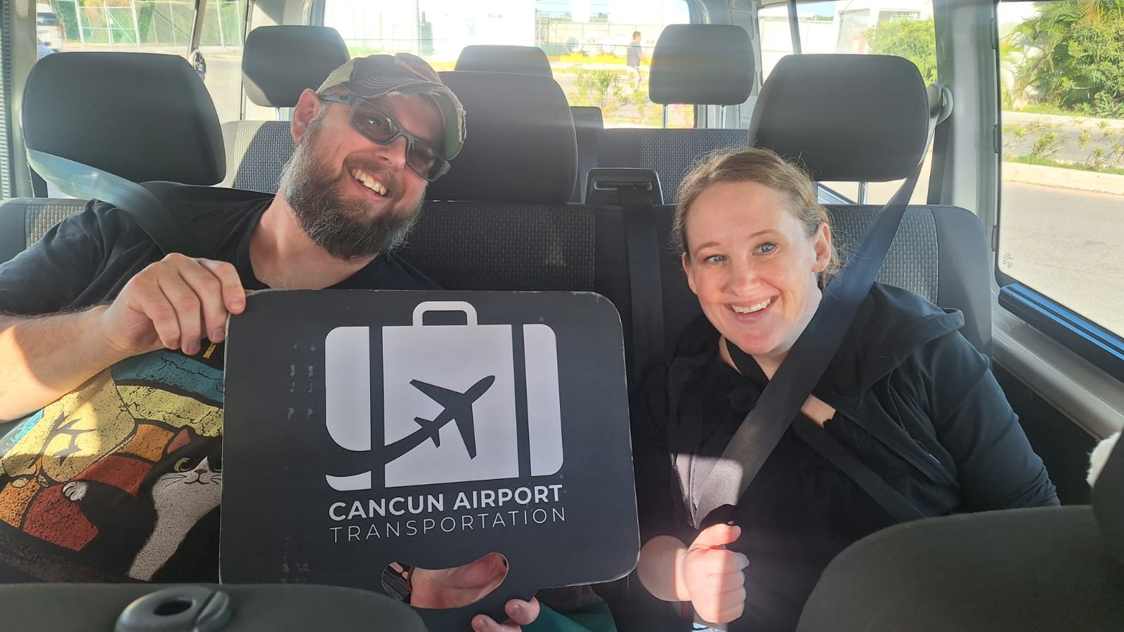Two smiling passengers wearing seatbelts sit in a van holding a black sign with a white luggage-and-airplane icon for airport transfers, sunlit interior with tropical greenery visible outside in Cancún.