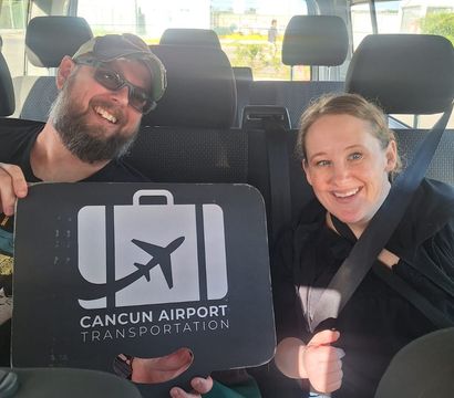 Two smiling passengers wearing seatbelts sit in a van holding a black sign with a white luggage-and-airplane icon for airport transfers, sunlit interior with tropical greenery visible outside in Cancún.