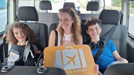 Three smiling passengers (an adult and two children) in an airport shuttle holding a yellow luggage sign with a suitcase and airplane icon
