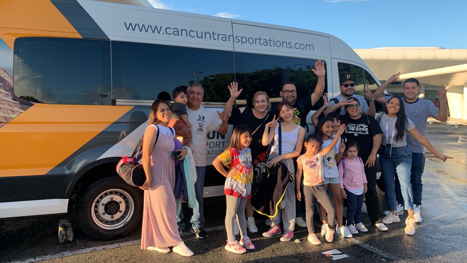 Smiling multigenerational family of travelers waving and posing beside a yellow-and-white airport shuttle van in sunny Cancun, kids with backpacks and luggage