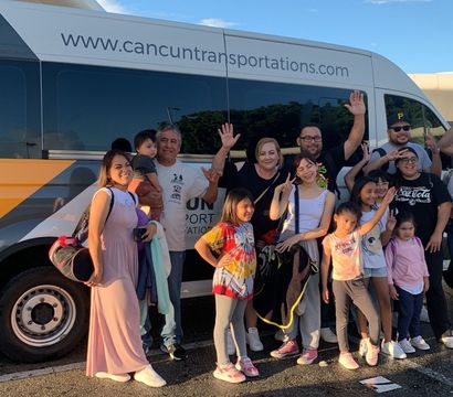 Smiling multigenerational family of travelers waving and posing beside a yellow-and-white airport shuttle van in sunny Cancun, kids with backpacks and luggage
