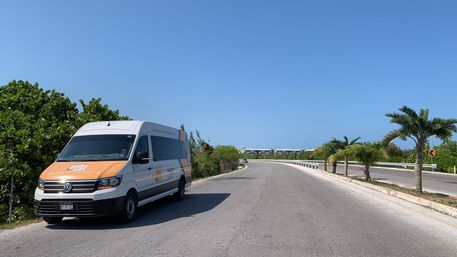 White-and-orange shuttle van parked on a sunny, palm-lined coastal road with clear blue sky and green foliage