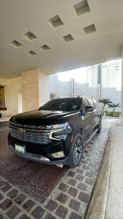 Black Chevrolet full-size SUV parked under a covered hotel porte-cochere with recessed square ceiling lights, cobblestone driveway and palm trees in the background.