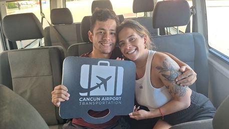 Two smiling travelers hugging inside a shuttle van holding an airport sign with an airplane icon, Cancun airport shuttle vibe