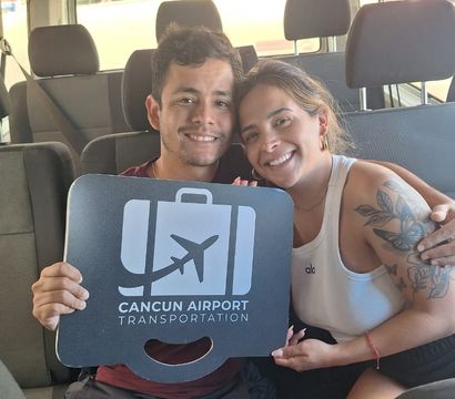 Two smiling travelers hugging inside a shuttle van holding an airport sign with an airplane icon, Cancun airport shuttle vibe