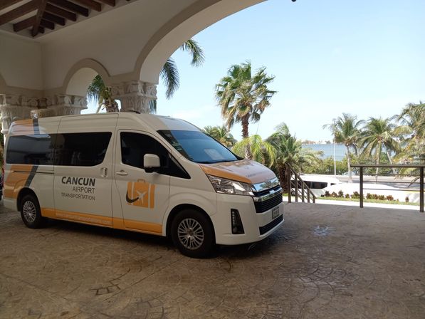 White-and-orange airport shuttle van parked under a resort portico with palm trees and turquoise sea in Cancún