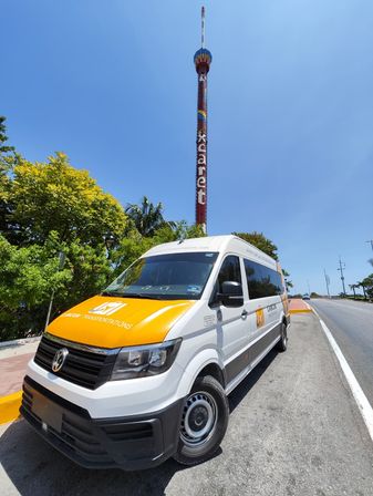 White-and-orange shuttle van parked on a sunny tropical coastal road beside a tall colorful amusement drop tower under a clear blue sky