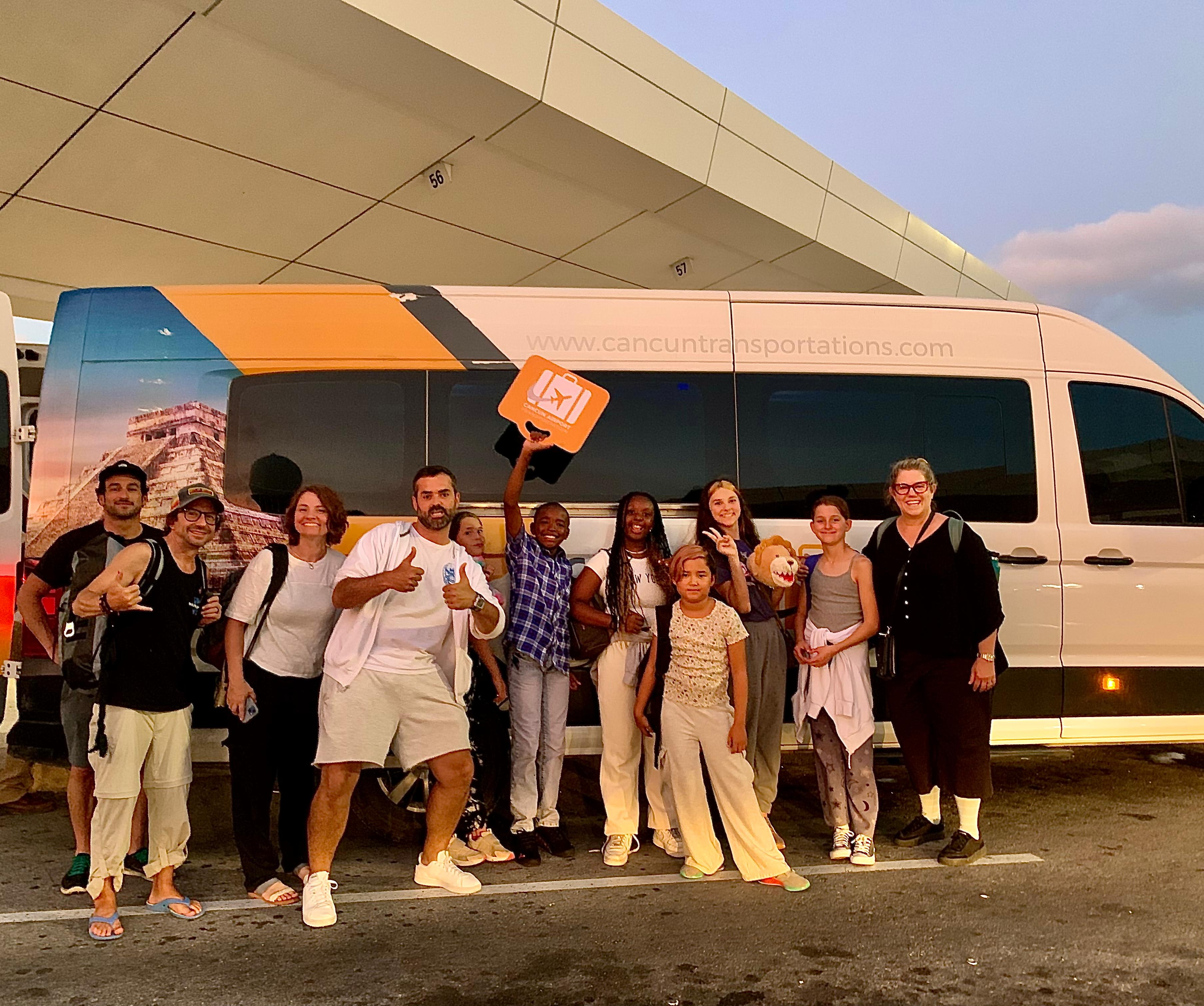 Smiling group of travelers, adults and children, posing at dusk in front of a white airport shuttle van on a tropical resort transfer, some giving thumbs-up and holding a luggage sign.