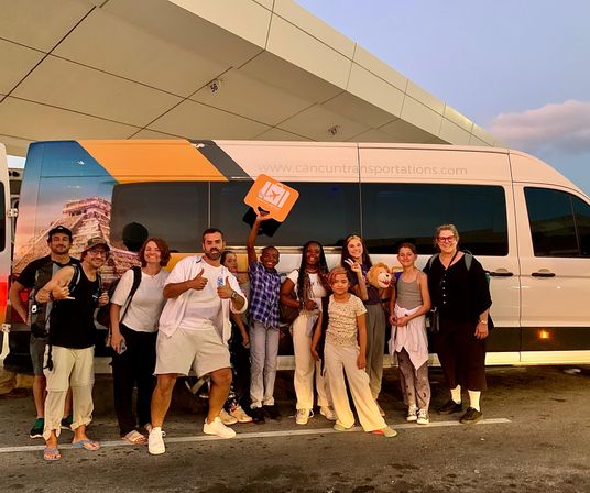 Smiling group of travelers, adults and children, posing at dusk in front of a white airport shuttle van on a tropical resort transfer, some giving thumbs-up and holding a luggage sign.