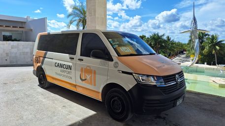 White-and-orange airport shuttle van parked at a sunny Cancun resort entrance with palm trees and a leaping marlin sculpture