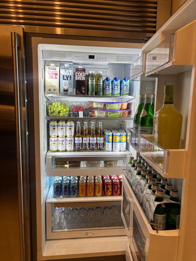 Open stainless-steel refrigerator in a modern kitchen, neatly stocked with milk and oat cartons, cold brew and juices, rows of bottled beer and canned seltzers, grapes and prepped meal containers.