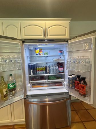 Open stainless-steel French-door refrigerator in a kitchen with white cabinets, neatly stocked with bottled water, canned drinks, juice, condiments and fresh produce.