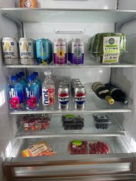 Open kitchen refrigerator with neatly arranged canned sparkling drinks and soda, bottled water and two wine bottles, packaged lettuce, and clamshells of strawberries, blueberries, raspberries and blackberries — colorful, organized snacks and beverages.