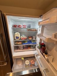 Open stainless-steel kitchen refrigerator showing milk jug, clear containers of strawberries and blueberries, eggs on a shelf, bags of vegetables in crisper drawers, condiments on the door, and two ice cream tubs in the bottom drawer.