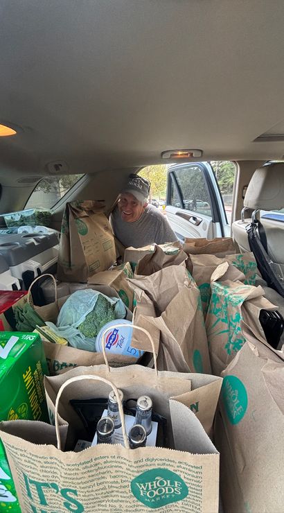 Smiling person leaning into the rear of an SUV packed with paper grocery bags and groceries — visible broccoli, yogurt tub, soda pack and bottled bottles through an open door.