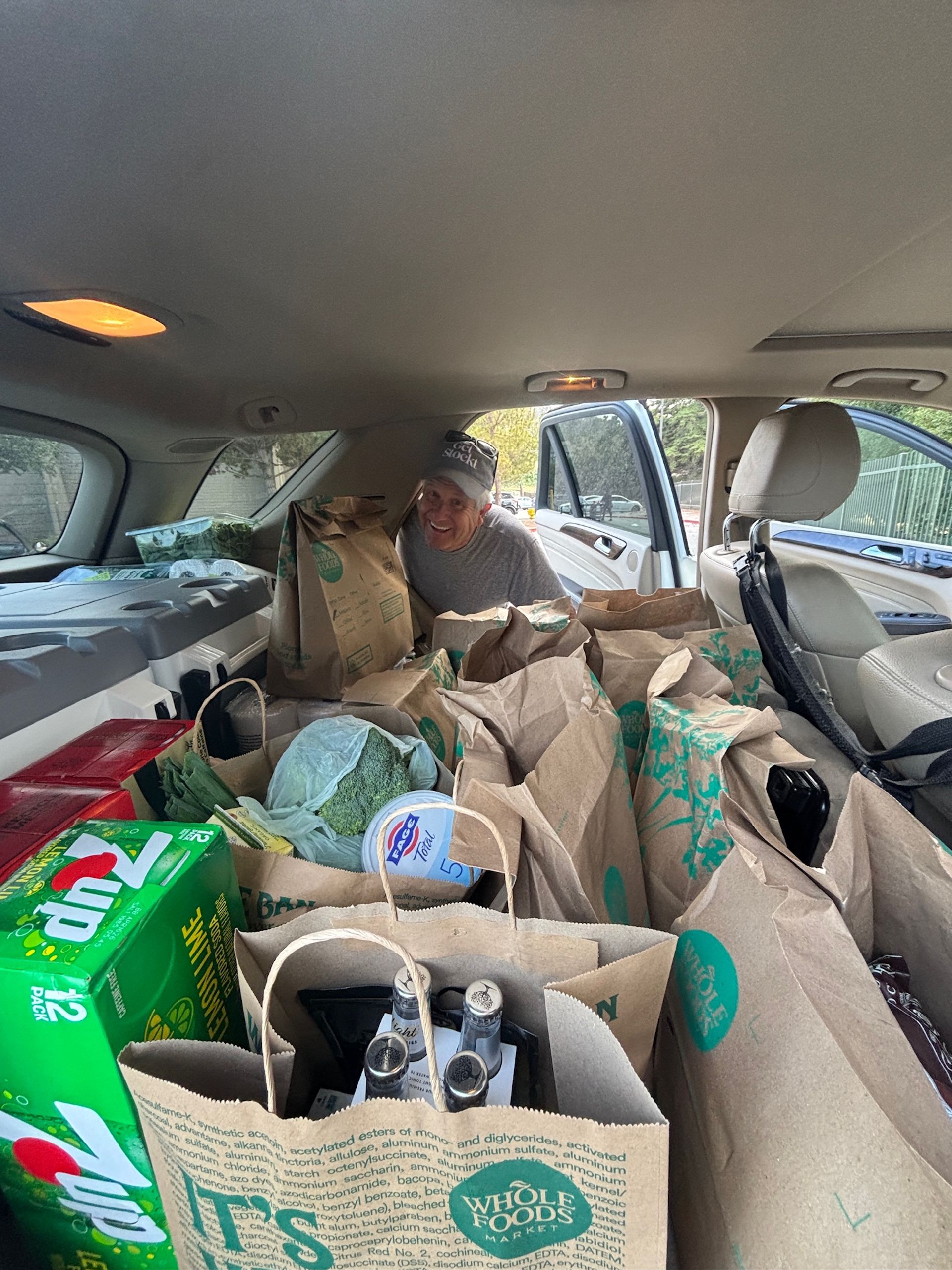 Smiling person leaning into the rear of an SUV packed with paper grocery bags and groceries — visible broccoli, yogurt tub, soda pack and bottled bottles through an open door.