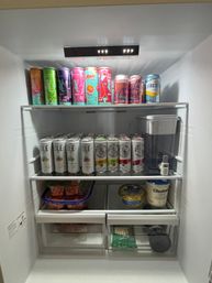 Open home refrigerator neatly stocked with bright slim canned drinks and sparkling waters on top shelves, hard seltzers beside a water‑filter pitcher, and containers of strawberries, yogurt and cheese in lower drawers.