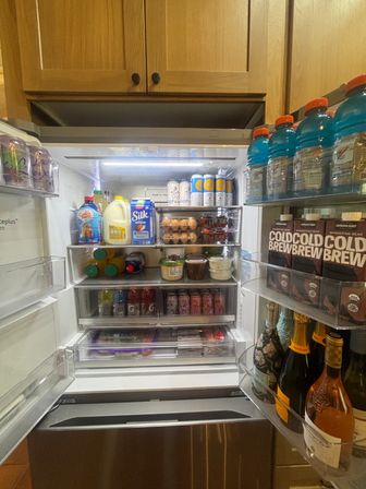Open stainless-steel kitchen refrigerator neatly stocked with milk and almond milk cartons, carton of eggs, jars and dips, rows of canned drinks, blue sports drinks and cold brew cartons in the door, and champagne bottles on the lower door shelf under wooden cabinets.