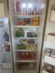 Open stainless-steel refrigerator neatly stocked with fresh groceries: apples, grapes, tomatoes, onions, egg cartons, cheese, yogurt, packaged meats and cans of sparkling drink.