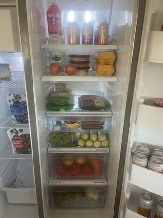 Open home refrigerator interior neatly stocked with fresh produce and groceries — apples, grapes, tomatoes, packaged cheese and yogurt, eggs, deli items on shelves and canned drinks in the door.