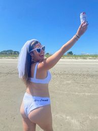 Smiling woman in a white bikini and bridal veil on a sunny sandy beach, wearing heart-shaped sunglasses and bride-themed earrings, raising a canned drink in celebration.