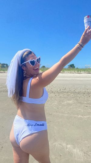 Smiling woman in a white bikini and bridal veil on a sunny sandy beach, wearing heart-shaped sunglasses and bride-themed earrings, raising a canned drink in celebration.