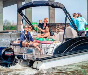 Group of friends cheering with drinks on a pontoon boat near a concrete bridge, colorful inflatable ring and outboard motor on a sunny summer river outing.