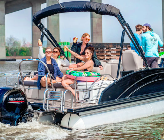 Group of friends cheering with drinks on a pontoon boat near a concrete bridge, colorful inflatable ring and outboard motor on a sunny summer river outing.