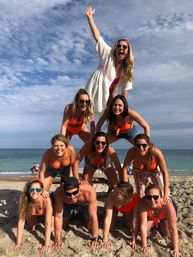 Group of ten friends forming a four-level human pyramid on a sunny sandy beach, wearing swimsuits and sunglasses with the ocean and blue sky in the background.