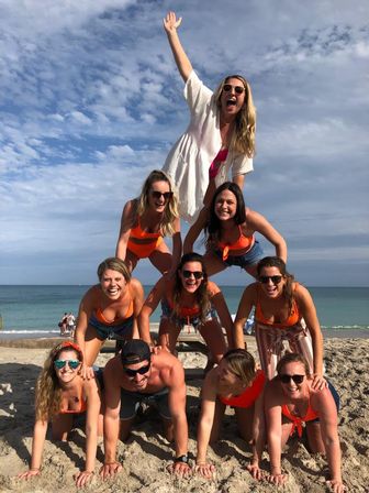 Group of ten friends forming a four-level human pyramid on a sunny sandy beach, wearing swimsuits and sunglasses with the ocean and blue sky in the background.