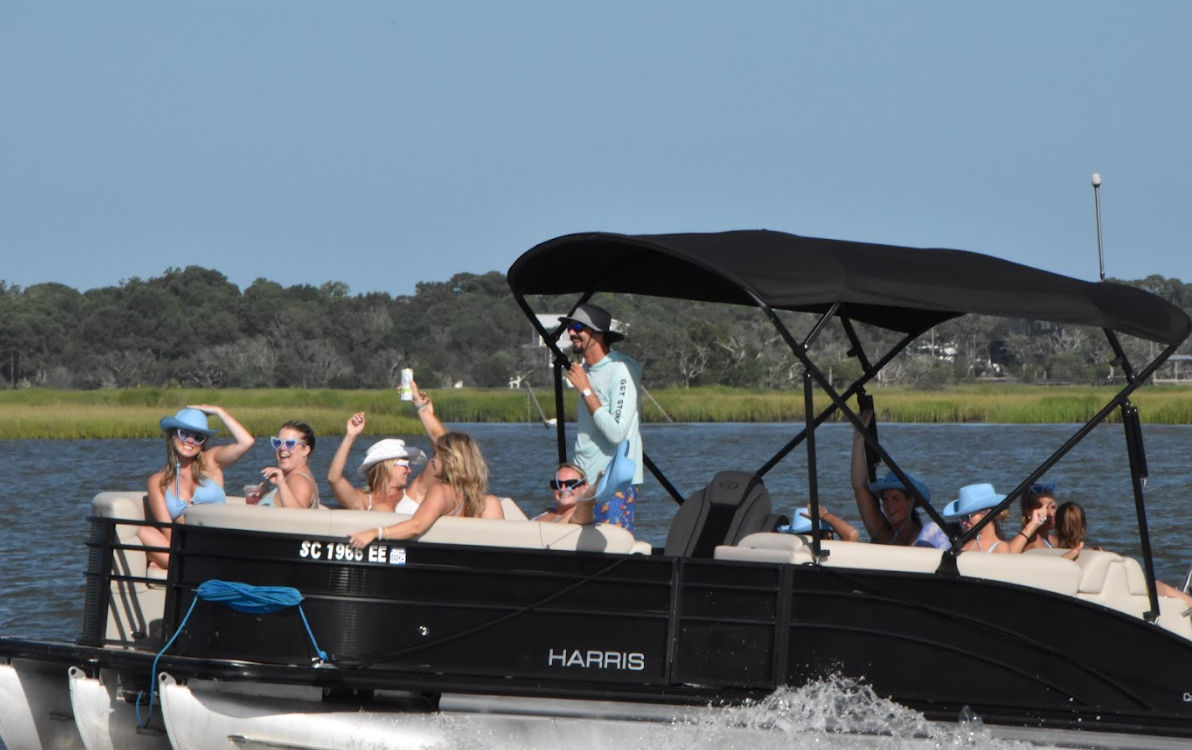 Group of friends on a black pontoon boat wearing sun hats and sunglasses, enjoying drinks while cruising past coastal marshland on a sunny summer day