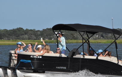 Group of friends on a black pontoon boat wearing sun hats and sunglasses, enjoying drinks while cruising past coastal marshland on a sunny summer day