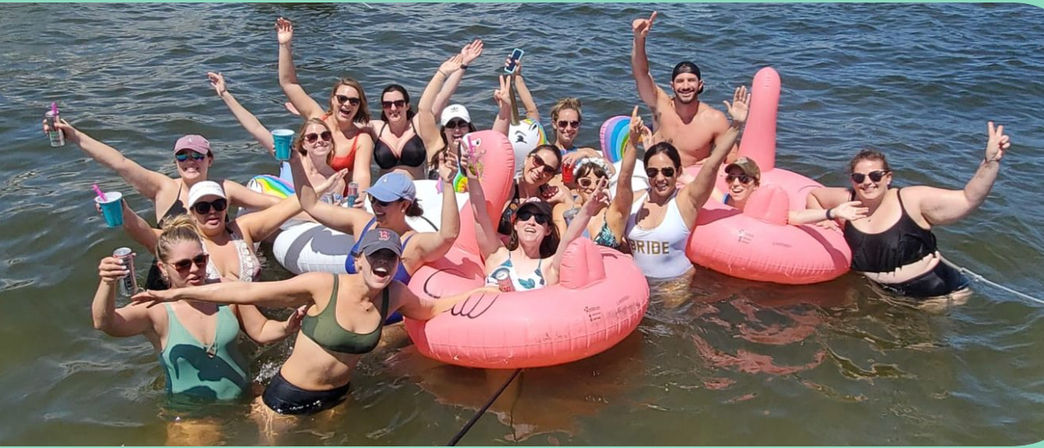 Sunny summer lake party with a large group on pink flamingo inflatables, holding drinks and cheering