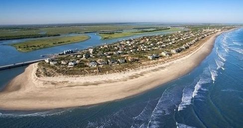 Aerial view of a narrow barrier island with beachfront houses, a wide sandy shore and breaking ocean waves on one side and calm inland waterways and marshes on the other.
